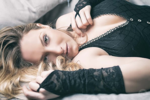 Portret van een vrouw liggend op bed in zwarte lingerie en kanten handschoenen, met haar blonde haar los om haar heen en haar blik rechtstreeks naar de camera.
