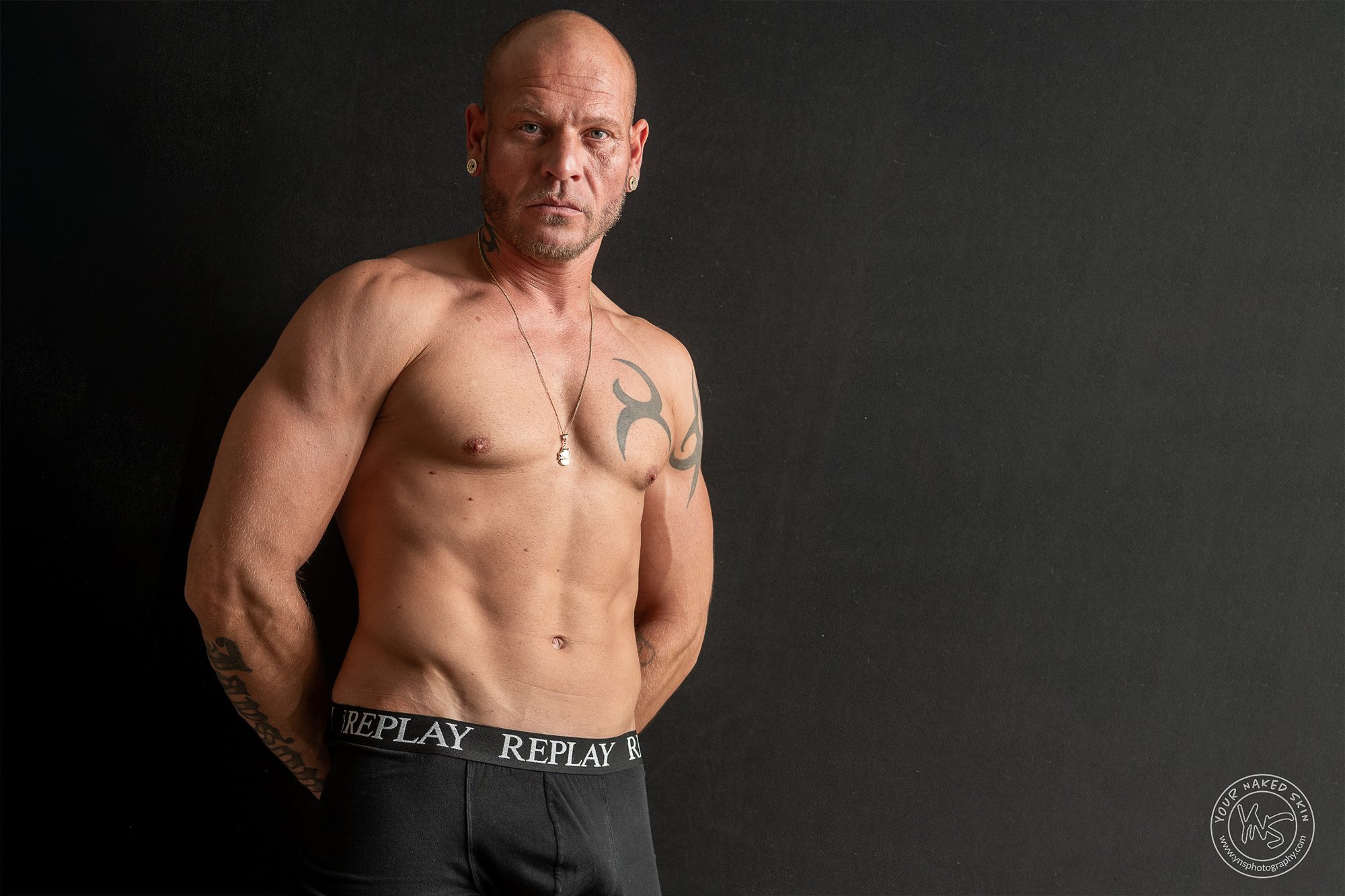 gespierde man met een tatoeage op zijn borst staat met ontbloot bovenlijf voor een donkere achtergrond.