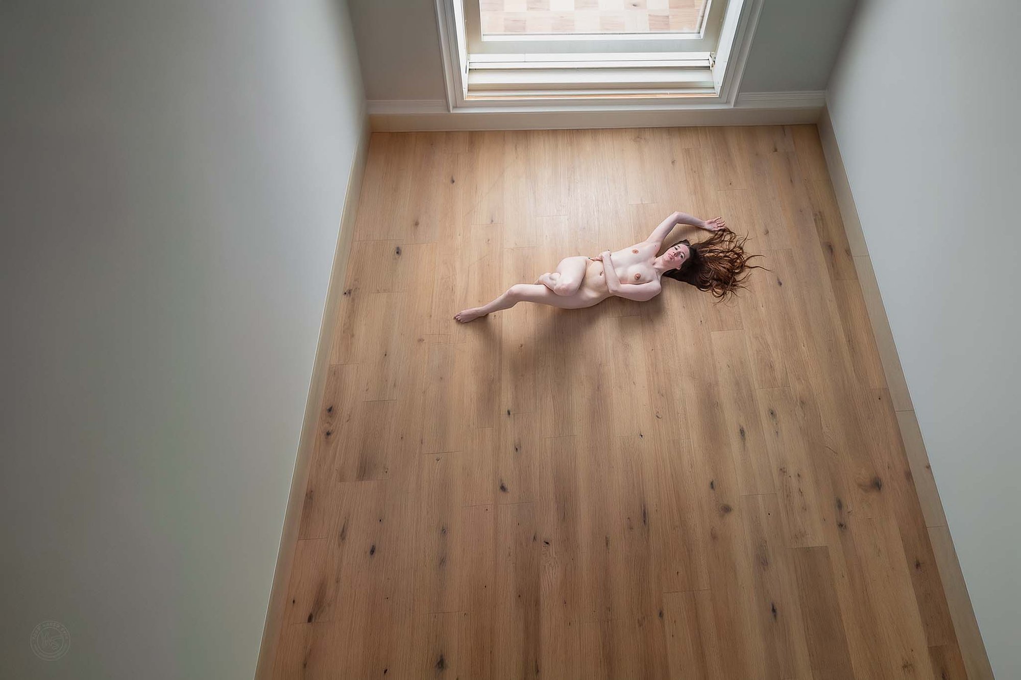 Bovenaanzicht van artistieke naaktfotografie van vrouw liggend op houten vloer in minimalistische ruimte met veel negatieve ruimte, focus op compositie, lijnen en natuurlijk licht.