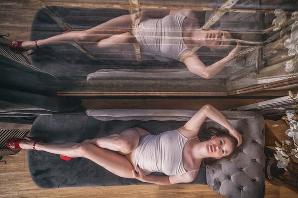 Vrouw in een witte top en rode hoge hakken ligt op een grijze chaise longue voor een grote spiegel, waarbij haar pose en reflectie samen een symmetrische compositie vormen.