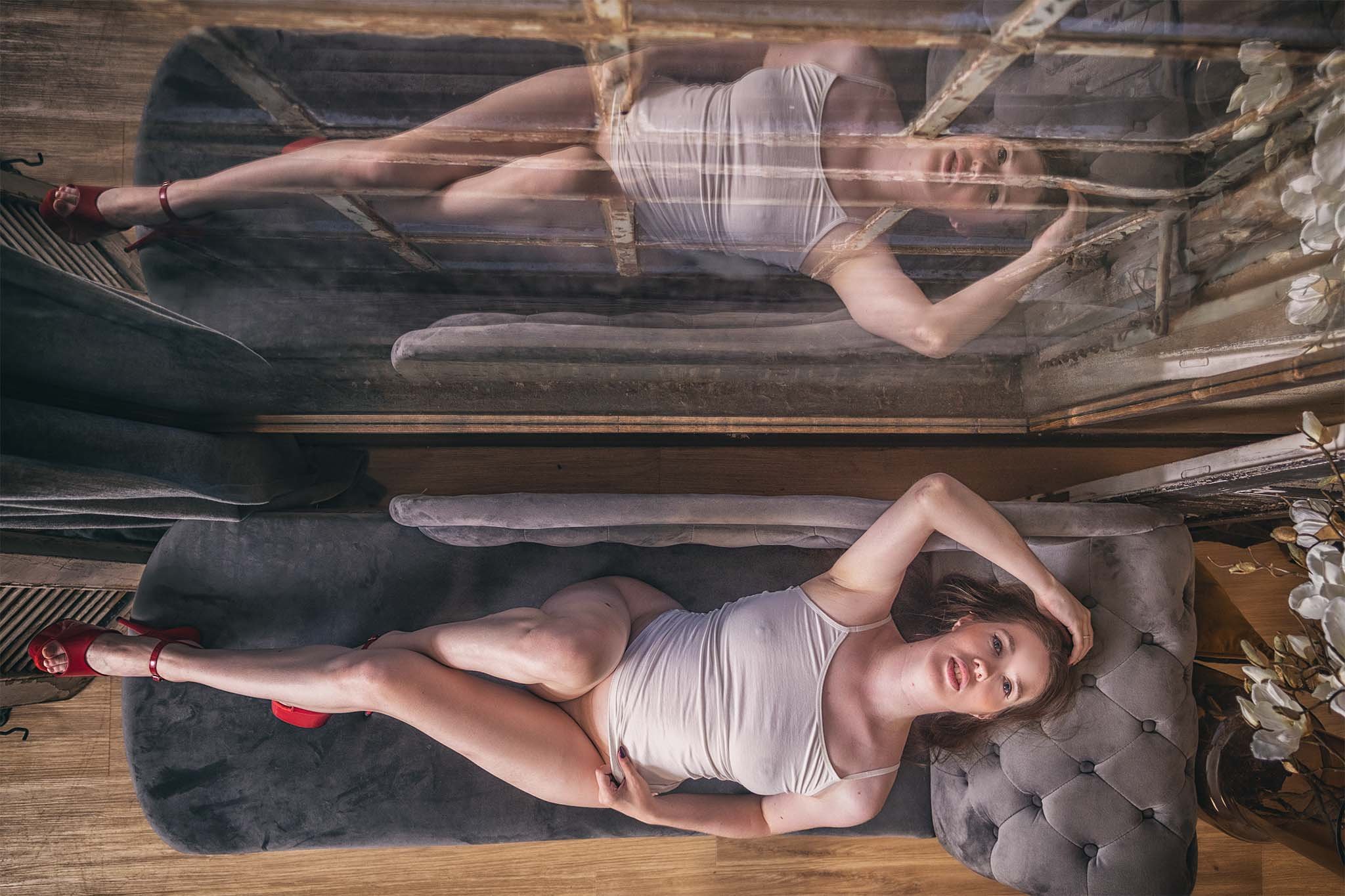 Vrouw in een witte top en rode hoge hakken ligt op een grijze chaise longue voor een grote spiegel, waarbij haar pose en reflectie samen een symmetrische compositie vormen.