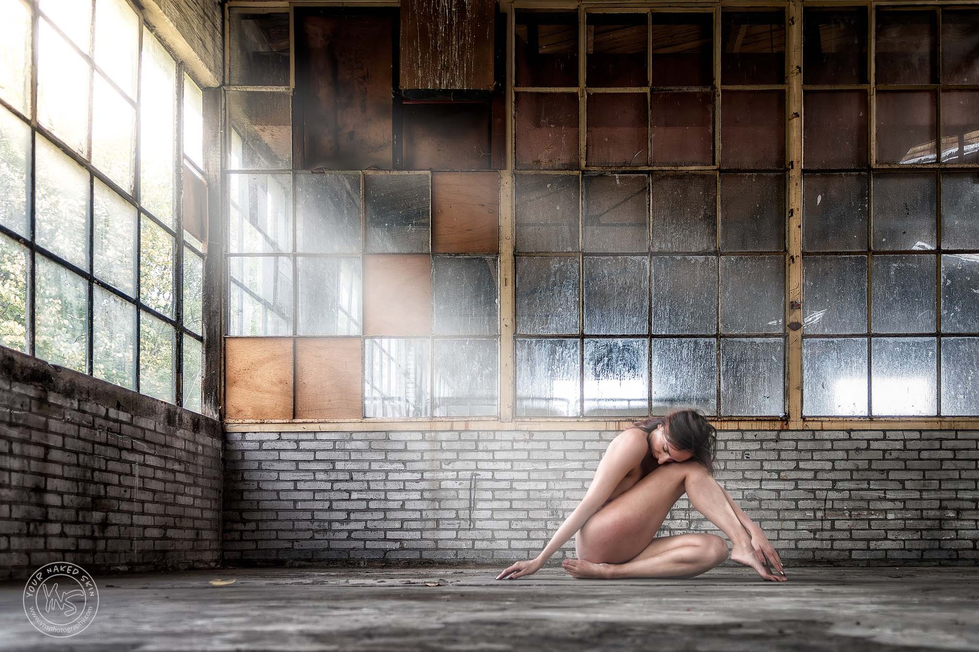Naakte vrouw zittend op de vloer in een industriële ruimte met grote ramen; natuurlijk licht valt zacht op haar lichaam en benadrukt houding, rust en vorm.