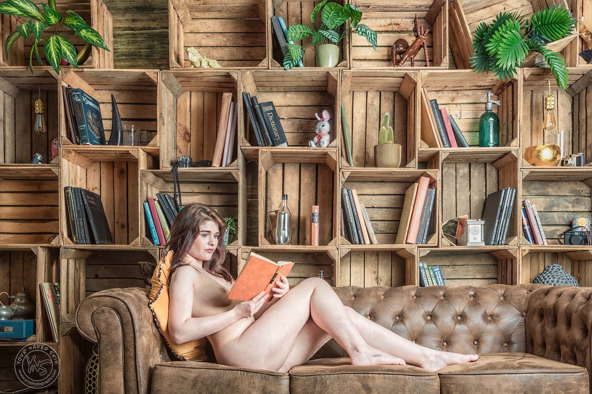 Artistieke naaktfoto van een vrouw die ontspannen op een leren bank ligt en een boek leest, gefotografeerd in een warme studio met houten kasten en boeken op de achtergrond.