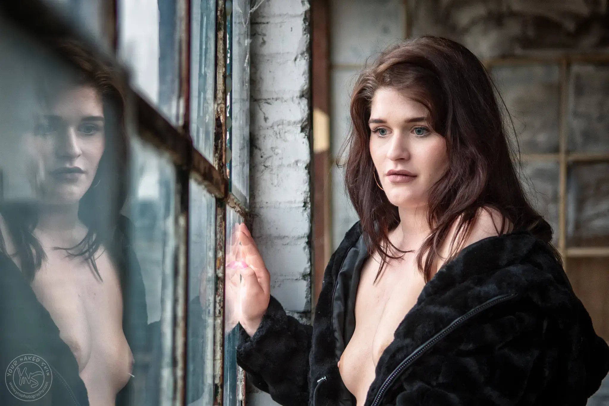 Een vrouw met donker haar staat bij een oud raam in een verlaten gebouw. Ze draagt een zwarte jas die van haar schouders glijdt en kijkt rustig naar buiten, terwijl haar reflectie zichtbaar is in het glas.