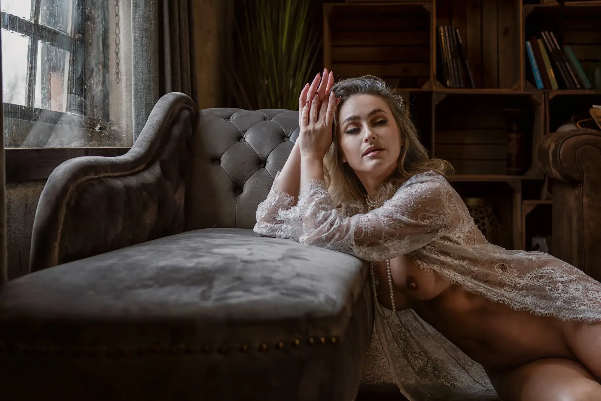 Zachte boudoir naaktfotografie van vrouw in kanten kimono bij raamlicht, sensuele pose naast fluwelen bank in warme sfeer — artistieke naaktfotografie door Your Naked Skin.