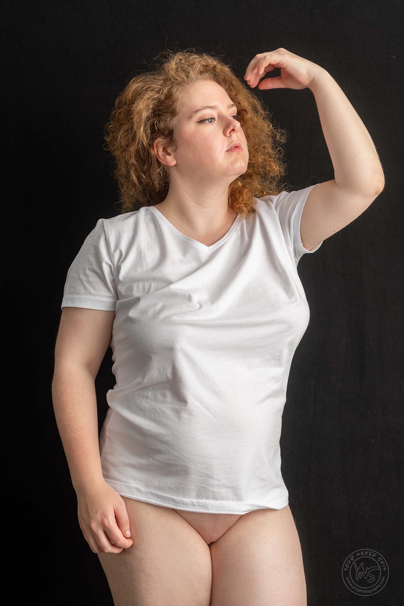 Vrouw met krullend rood haar draagt een wit T-shirt en staat tegen een zwarte achtergrond, met haar arm omhoog en haar blik zijwaarts gericht, gefotografeerd in een rustige studio-opstelling met zacht licht.