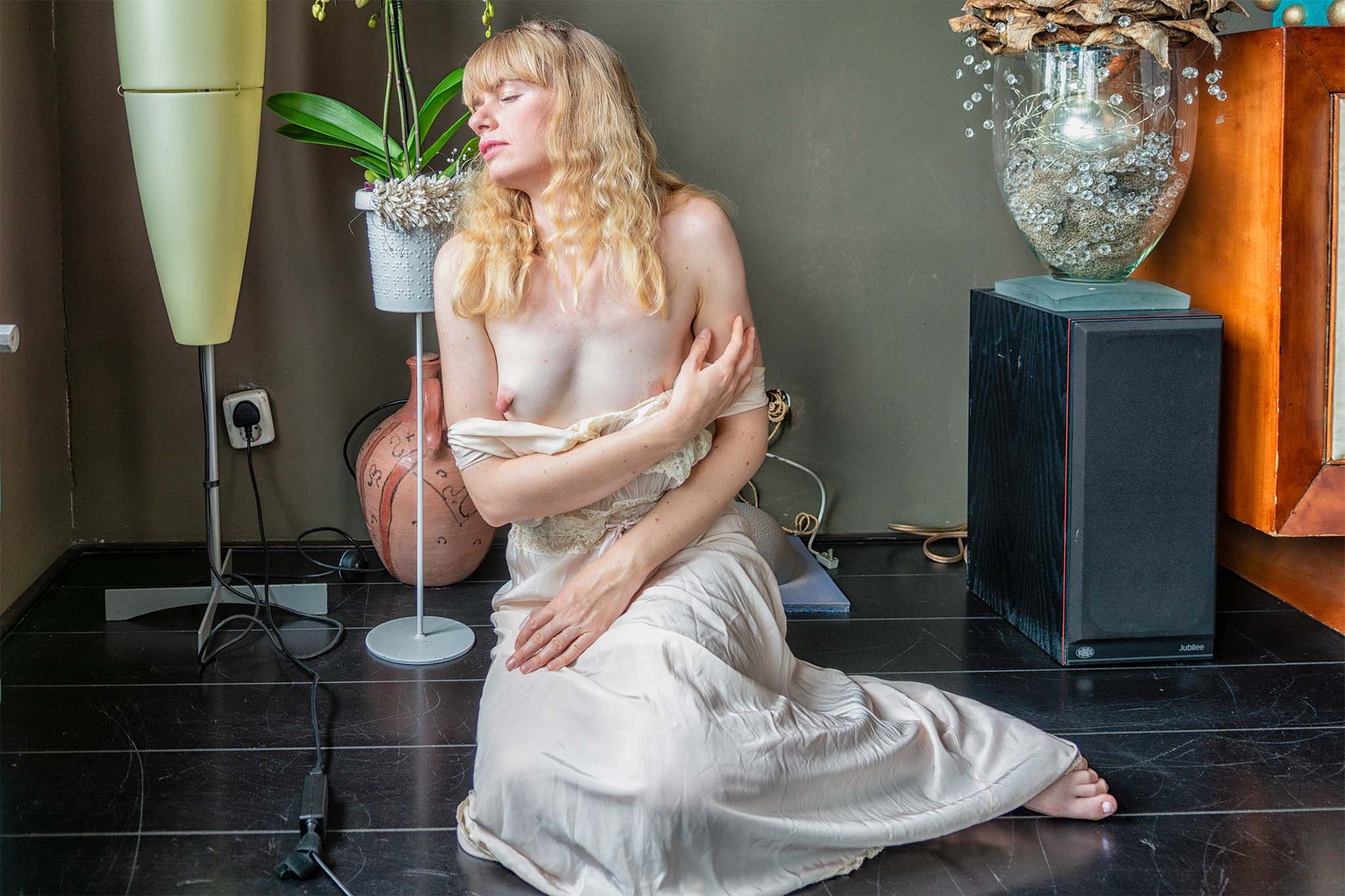 Fine art naaktfoto van blonde vrouw gehuld in zijdeachtige stof, zittend op donkere vloer bij zacht daglicht, met intieme en schilderachtige uitstraling.