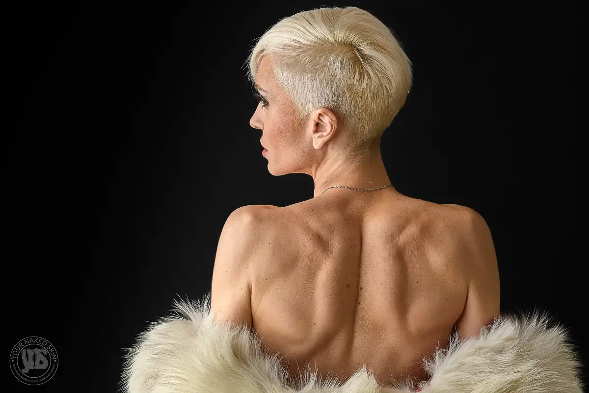 Naakte vrouw met kort, blond pixiekapsel poseert met haar rug naar de camera tegen een zwarte achtergrond. Ze draagt een lichte bontachtige stola over haar armen. De belichting benadrukt de natuurlijke vormen van haar rug en schouders, wat een elegante en tijdloze sfeer creëert.