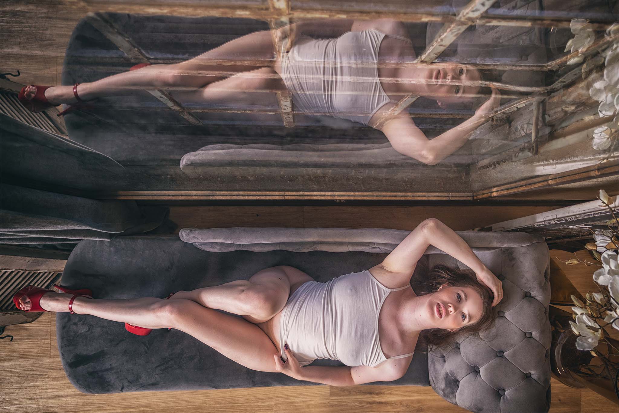 Vrouw in een witte top en rode hoge hakken ligt op een grijze chaise longue voor een grote spiegel, waarbij haar pose en reflectie samen een symmetrische compositie vormen.