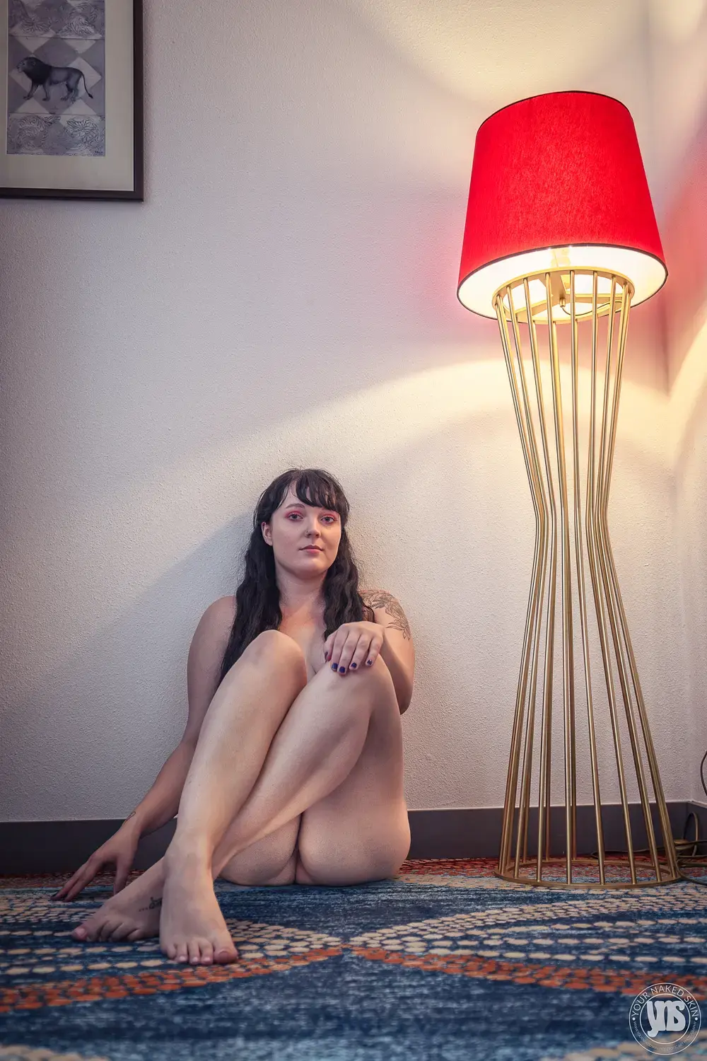 Naakte vrouw zittend op de grond in een hoek, benen gekruist voor haar lichaam, met één hand op de knie en de andere op de vloer – een natuurlijke en zelfverzekerde pose in warme sfeerverlichting.